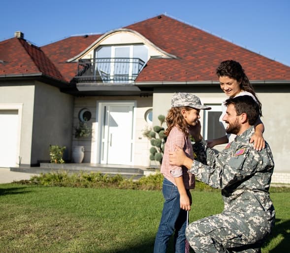 A veteran in uniform kneeling to embrace his daughter in front of a home